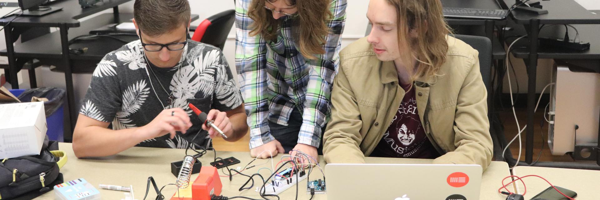 Three students working on creating a project for a class using a soldering kit and other tools.