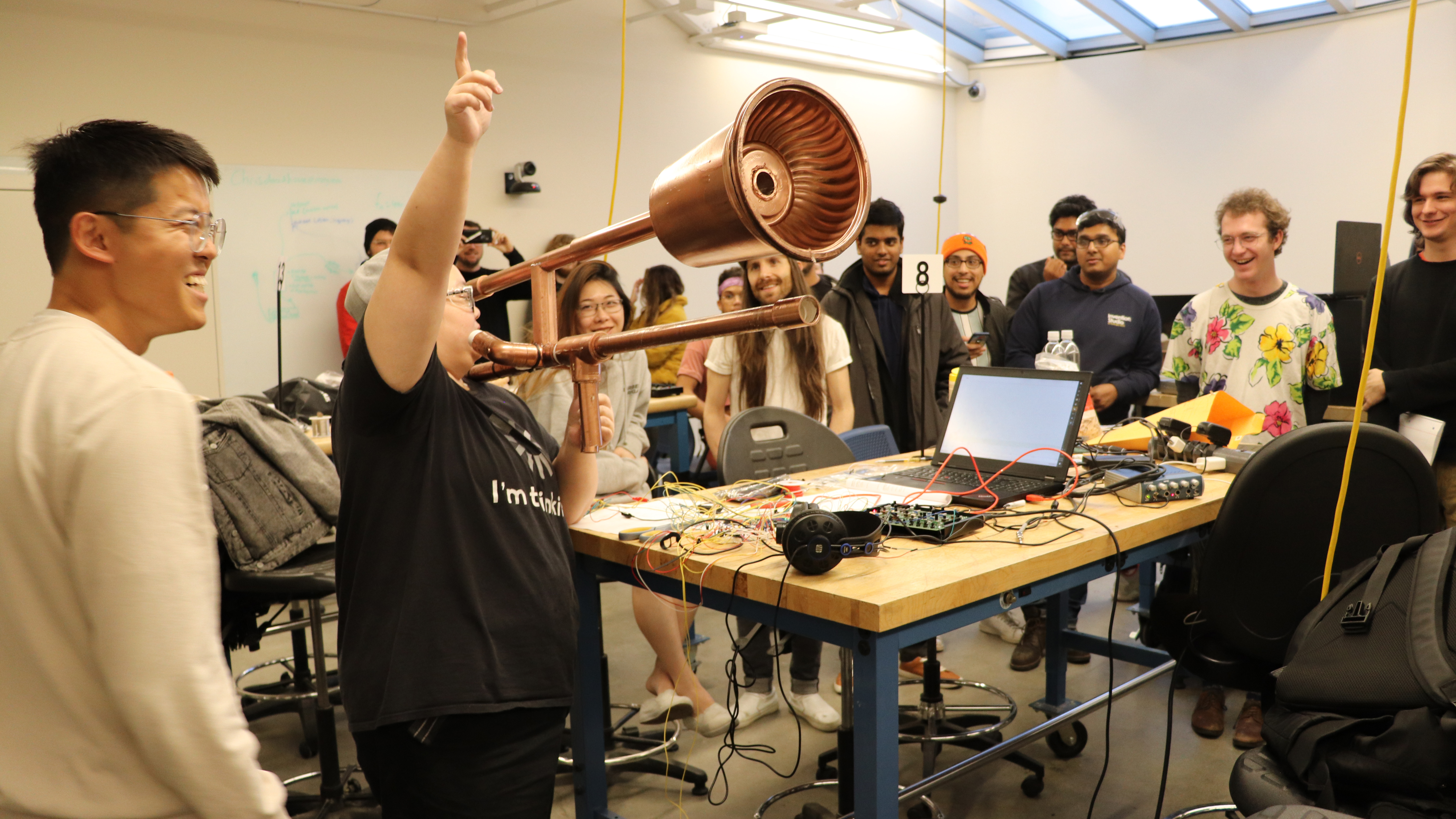 A Hackathon competitor plays a modified trombone while other smiling competitors watch.