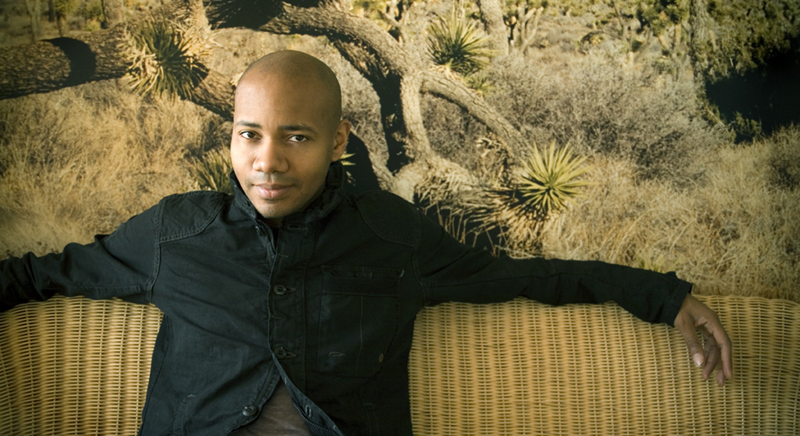 DJ Spooky wearing a dark jacket while lounging on a light tan couch.