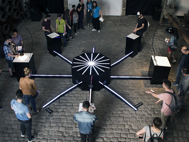 A standing display with a circle in the middle with interfaces in a pattern surrounding the center display, with people around the display.