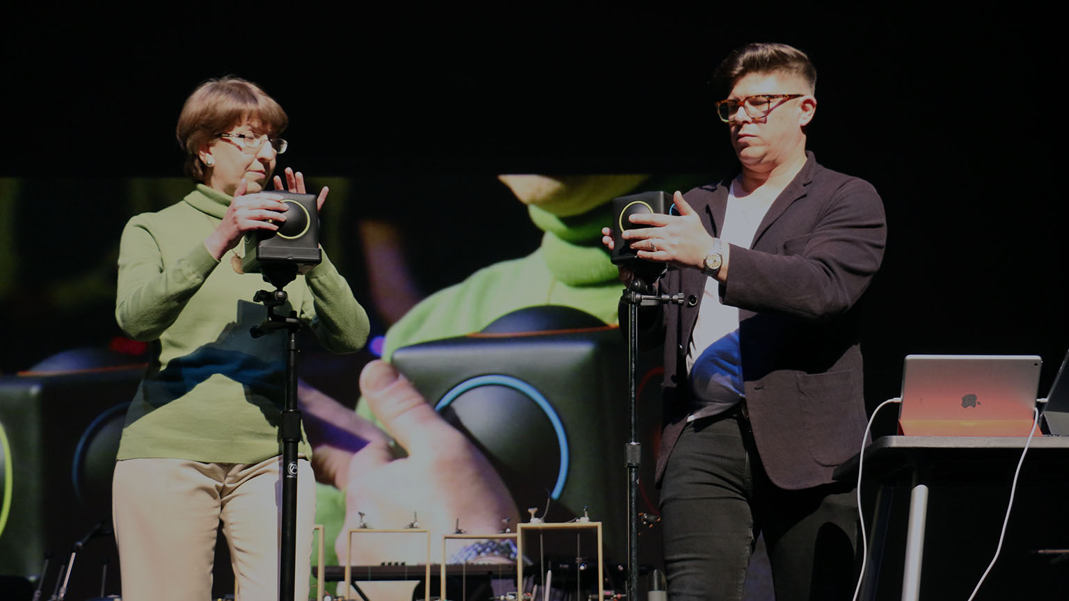 Two people on stage using a cube with different colored sides to play music.
