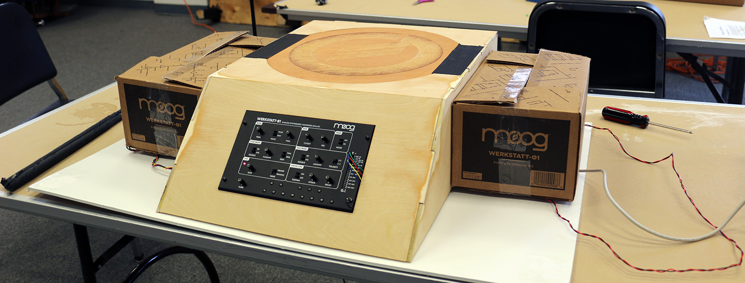 A wooden drum with a Moog Werkstatt in the middle, with two cardboard boxes that are also playable on either side.
